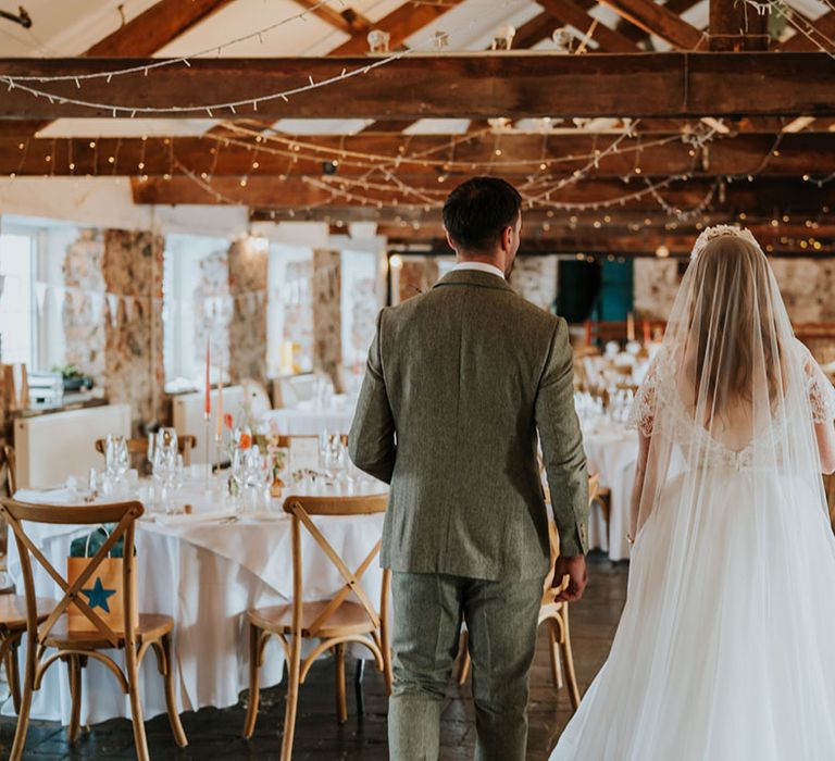 The groom in a tweed suit walks into the reception room with the bride in lace and tulle wedding dress 