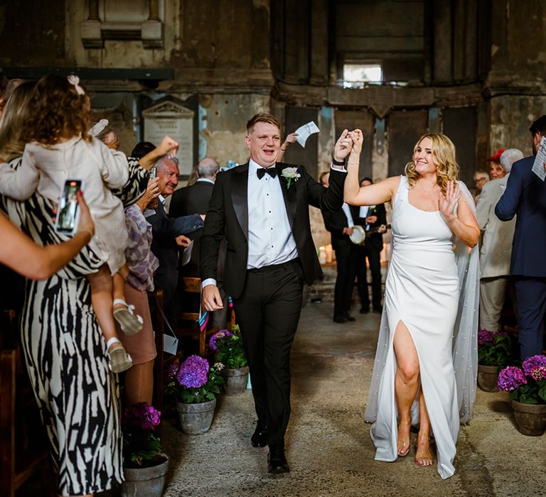 Bride in fitted wedding dress with pearl embellished cape walks with her groom in black tie after wedding ceremony 