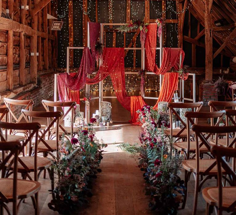 Brides standing at the end of the alter at Silchester Farm with bold crushed velvet fabrics, pheasant feathers, silver birch log stacks, candles, mismatched gold candlesticks, dried autumnal floral arrangements, red wedding drapes, fairy lights, festoon chandeliers and a string trio