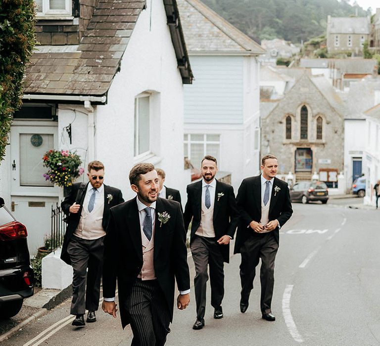 Groom & his groomsmen walk together wearing morning suits, blue ties and floral buttonholes 