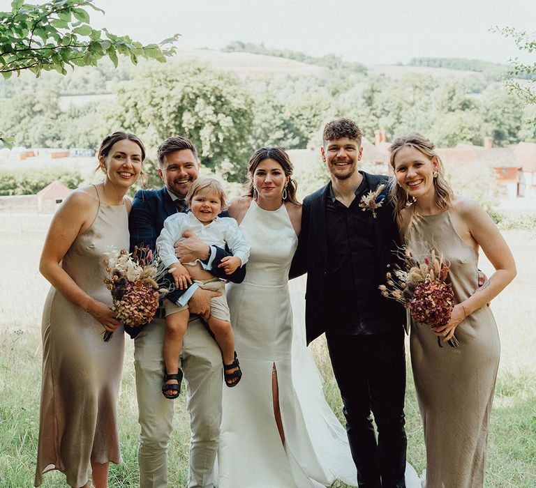 Bridesmaids in satin champagne gold bridesmaid dresses and bride in front flit dress, groom in all black suit and their son for the wedding baptism 