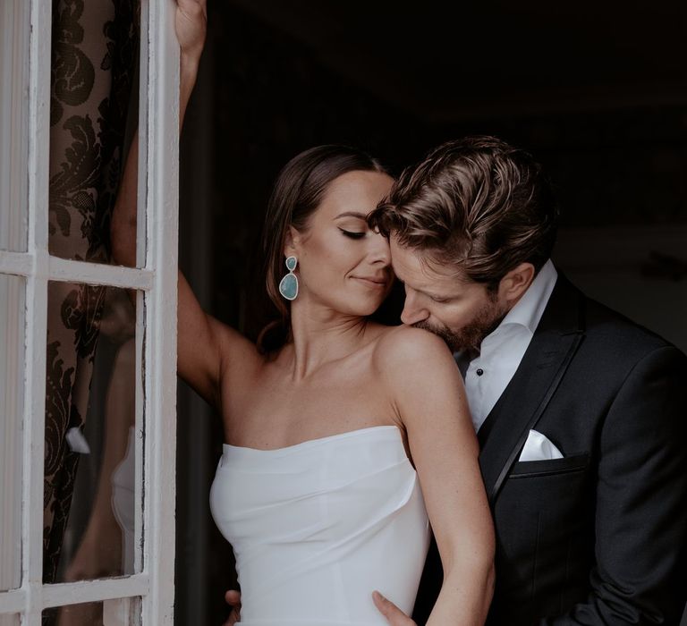 Bride in satin strapless wedding dress and azure blue drop earrings being kissed on the shoulder by groom in classic black tux with white pocket square 