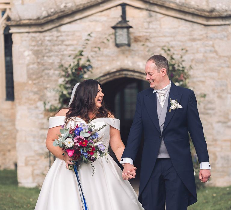 Bride in an off the shoulder pleated skirt wedding dress holding a purple and blue wedding bouquet with the groom in a navy morning suit with white rose buttonhole 