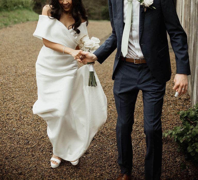 Bride in off the shoulder Eva Lendel wedding dress walking with the groom in a navy suit and sage green tie for their classic rustic luxe wedding 