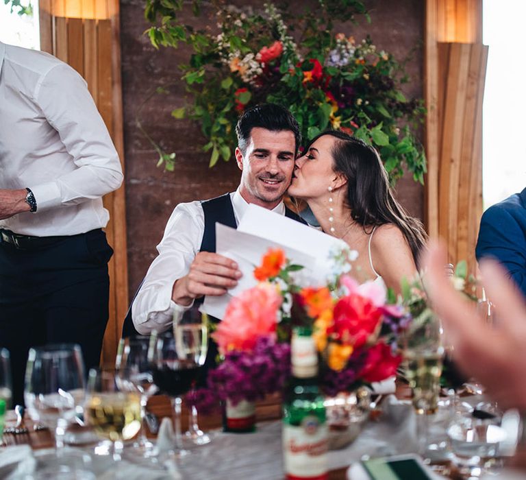 The bride wearing long statement pearl earrings leans in and kisses the groom on the cheek as he prepares to read his wedding speech 