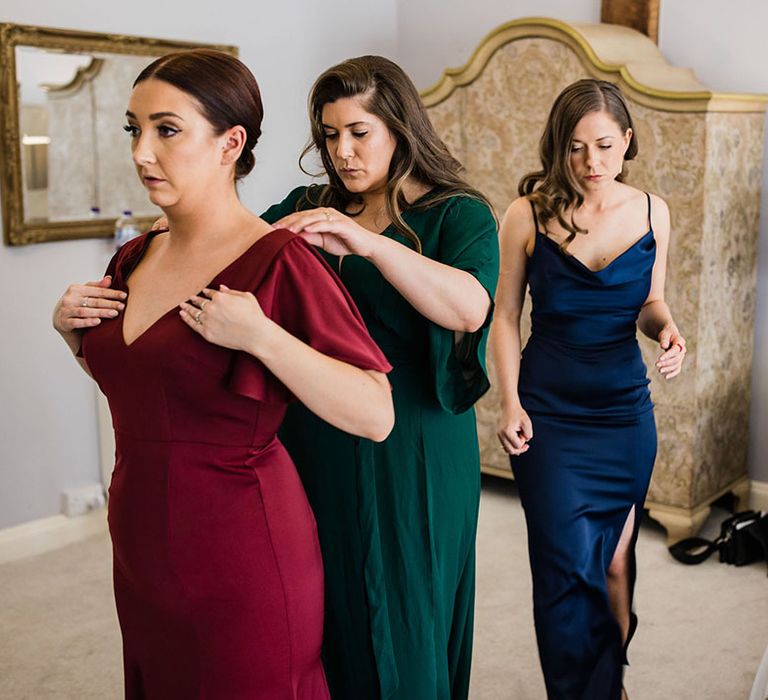 The bridesmaids help each other get ready all wearing jewel tone mismatched bridesmaid dresses 