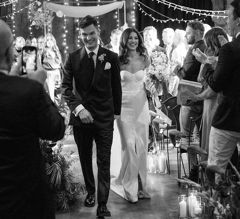 The bride and groom walk down the aisle at the rustic Tythe Barn wedding venue with white flowers 