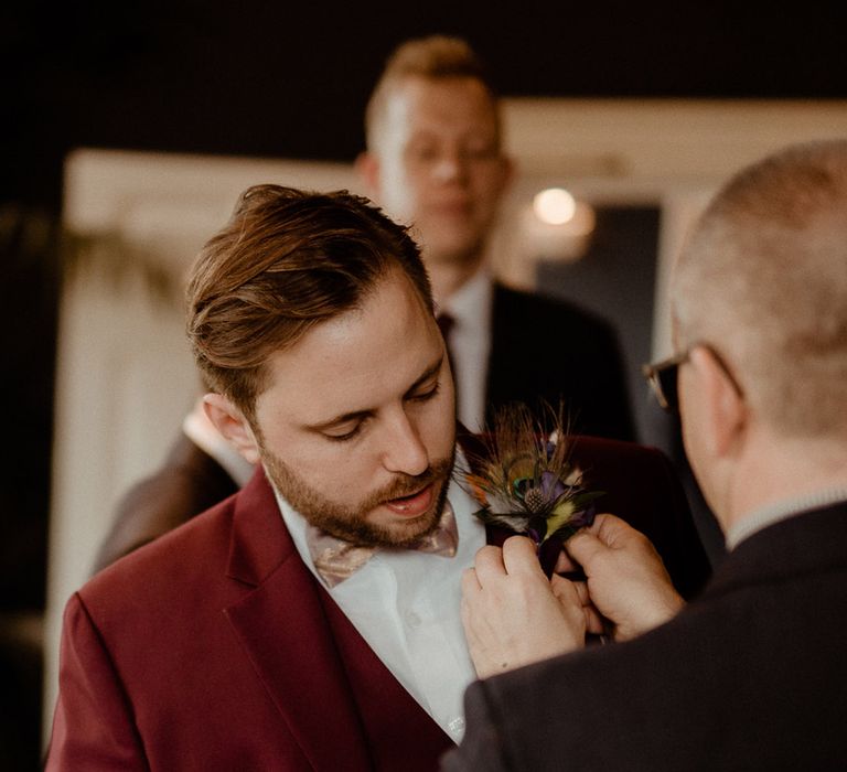 A groomsman helps put on the groom's dried flower peacock feather buttonhole to match the boho Newton Hall wedding in a burgundy three piece suit 