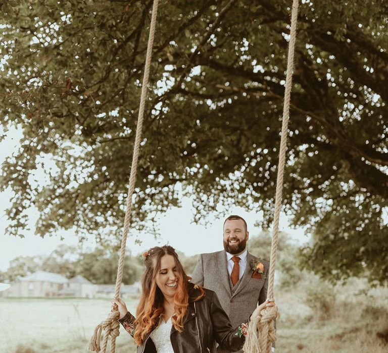 Bride in lace wedding dress wearing wedding leather jacket on a swing being pushed by groom in light grey suit 