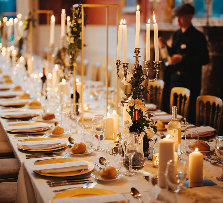 Grittleton House reception room with classic wedding tablescape including white table cloth, tapered candles in gold candelabras, pillar candles, gold frame decorations, gold plates, gold cutlery and dried flower and white rose floral arrangements 