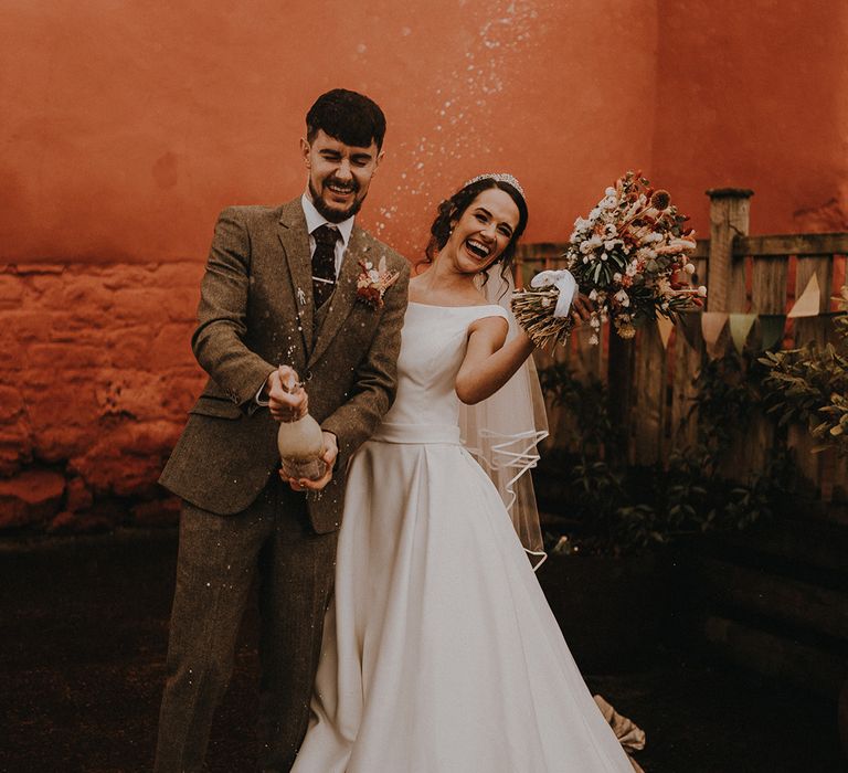 The groom in a grey three piece suit pops champagne with the bride in an off the shoulder wedding dress for fun wedding celebrations 