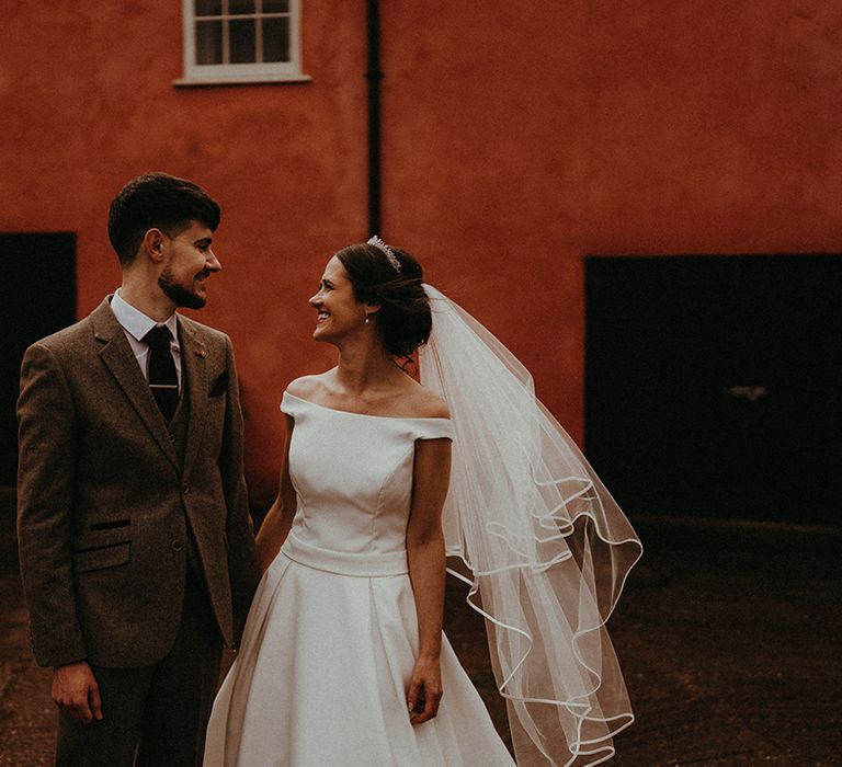 Groom in grey three piece suit with red tie and dried flower buttonhole poses with the bride in an off the shoulder wedding dress for rustic barn wedding 