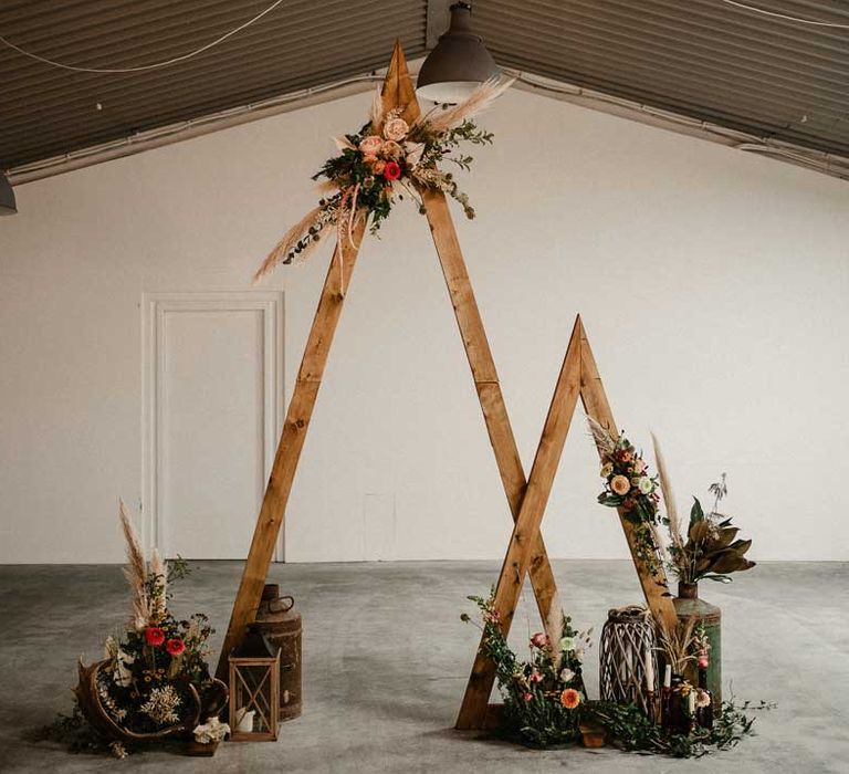 One small and one large wooden triangular wedding arch with pampas grass, foliage, and garden rose arrangements attached, rustic wedding decorations, large plants in vases and pillar candles 