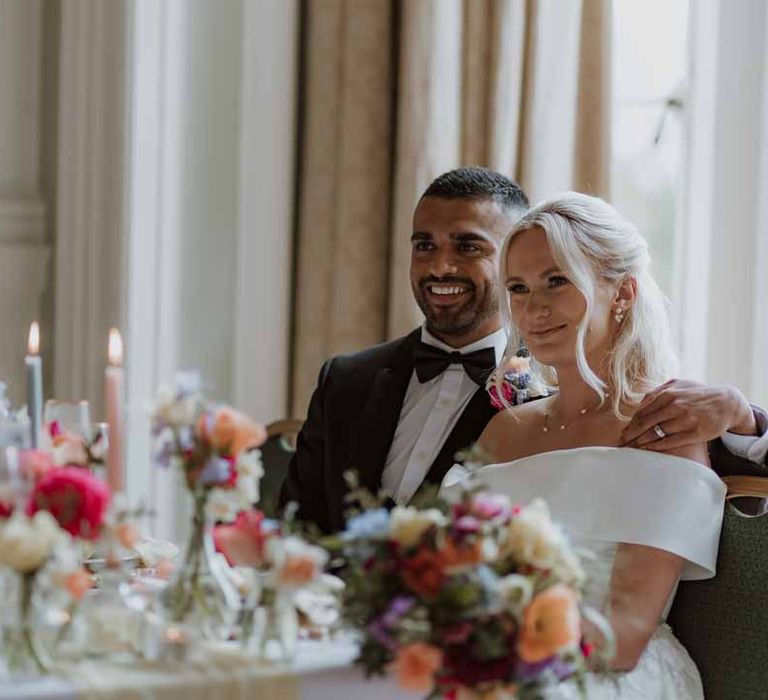 Bride in off the shoulder lace wedding dress and groom in classic three piece grooms tuxedo with colourful spring boutonniere sitting by spring themed wedding tablescape with orange poppies, pink and red roses, jasmine and bluebells on white wedding tablecloth and off-white satin table runner with pastel tapered candles, gold-rimmed wine glasses and classic crockery 