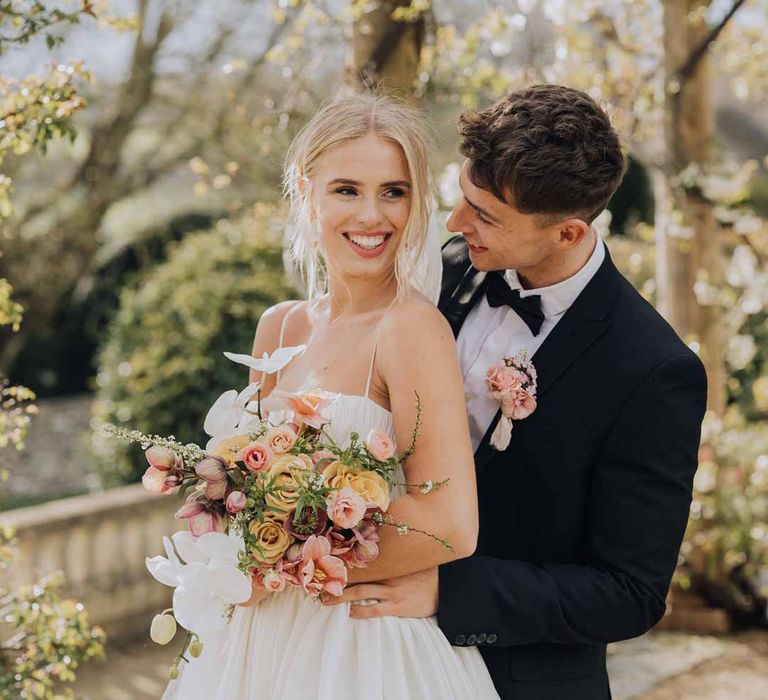 Bride in Watters strappy sleeveless princess wedding dress and chapel length wedding veil holding blush toned bridal bouquet with poppies, ranunculus, roses, tulips and foliage posing with groom in classic black tuxedo with bowtie and blush rose boutonniere 