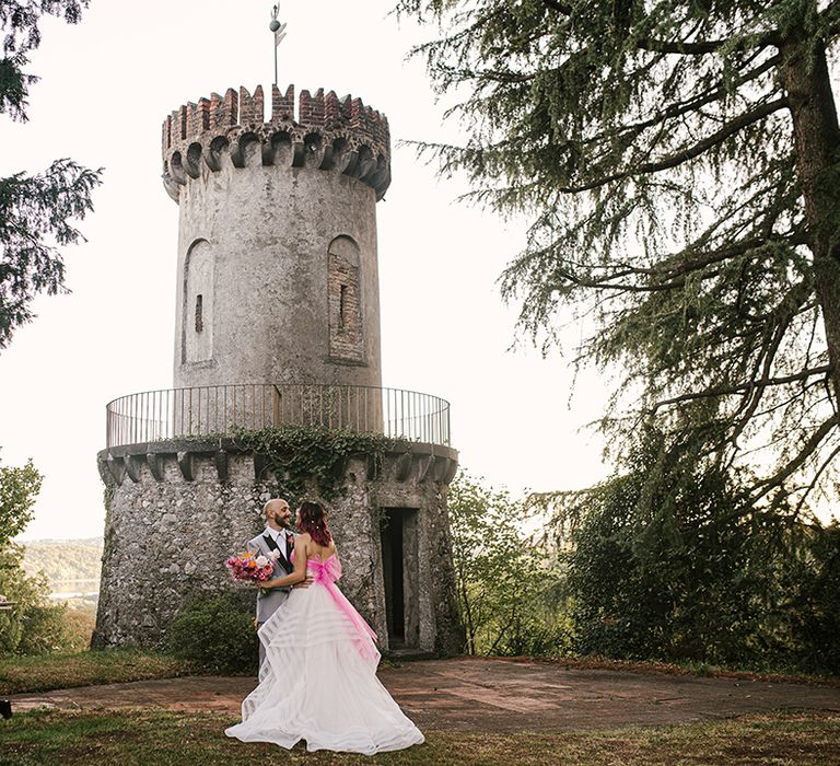 groom in a grey suit embracing his bride in a strapless wedding dress with pink bow standing in front of a castle