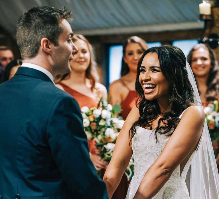 Bride in a-line tulle wedding dress with beaded lace applique with petals and a square neckline with side tulle cutouts and church-length veil holding hands with groom at the alter wearing navy grooms morning suit 