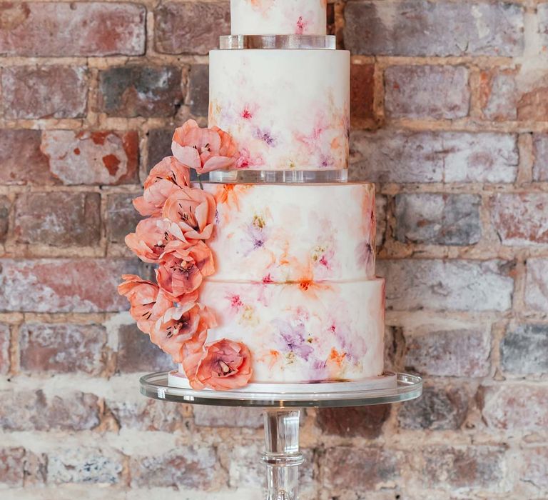 Four tiered hand-painted iced wedding cake with hand-made edible flower decorations on clear cake stand at Rackleys Barn