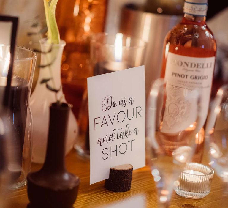 Minimalist white wedding sign "Do us a favour and take a shot" with wedding drinks behind it at Kingscote Barn Tetbury 