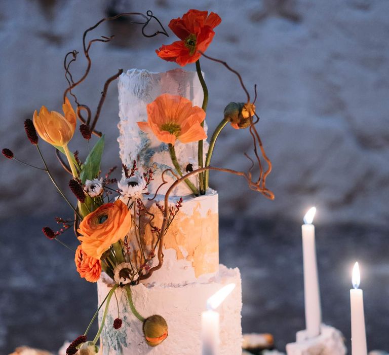 Three tiered iced wedding cake with intricate rose and poppy wedding cake topper on wedding grazing table surrounded by tapered candles in Margate Caves