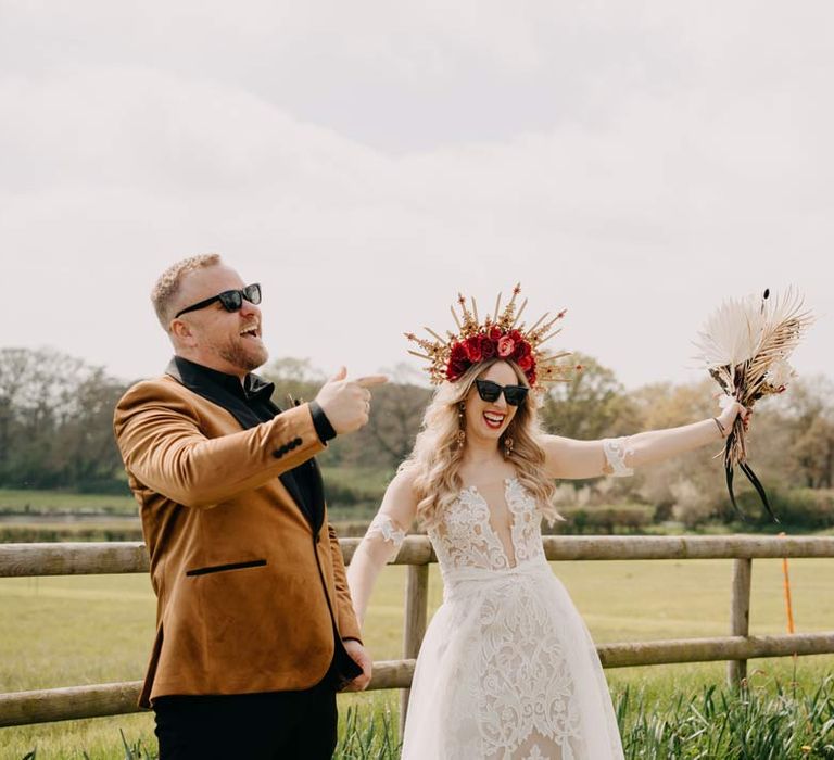 Bride in off shoulder lace wedding dress with corset style top and red rose, gold halo bridal crown and sunglasses holding red rose, pampas grass and dried flower bridal bouquet walking with groom incrushed velvet burnt orange grooms blazer and dried flower boutonniere and sunglasses at Southlands Barn wedding