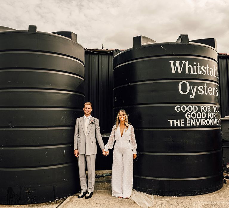 Groom in a grey suit holding hands with his bride in a wide leg bridal jumpsuit at East Quay Whitstable wedding venue 