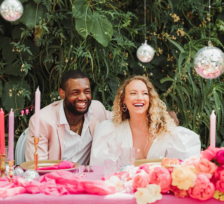 Hanging disco ball decorations with bride and groom sitting at hot pink sweetheart table with groom in pink suit and bride in puff sleeve wedding dress