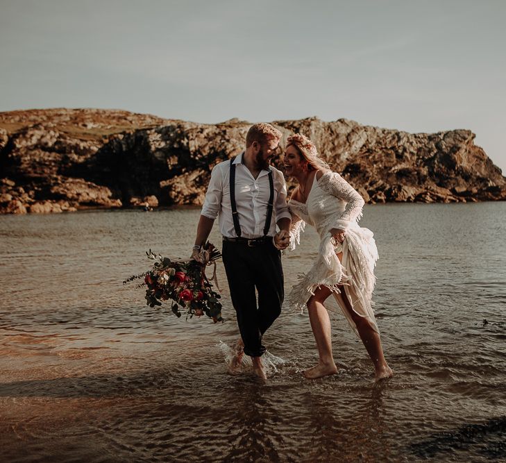 Groom in shirt and braces and bride in lace wedding dress share laughter in the sea