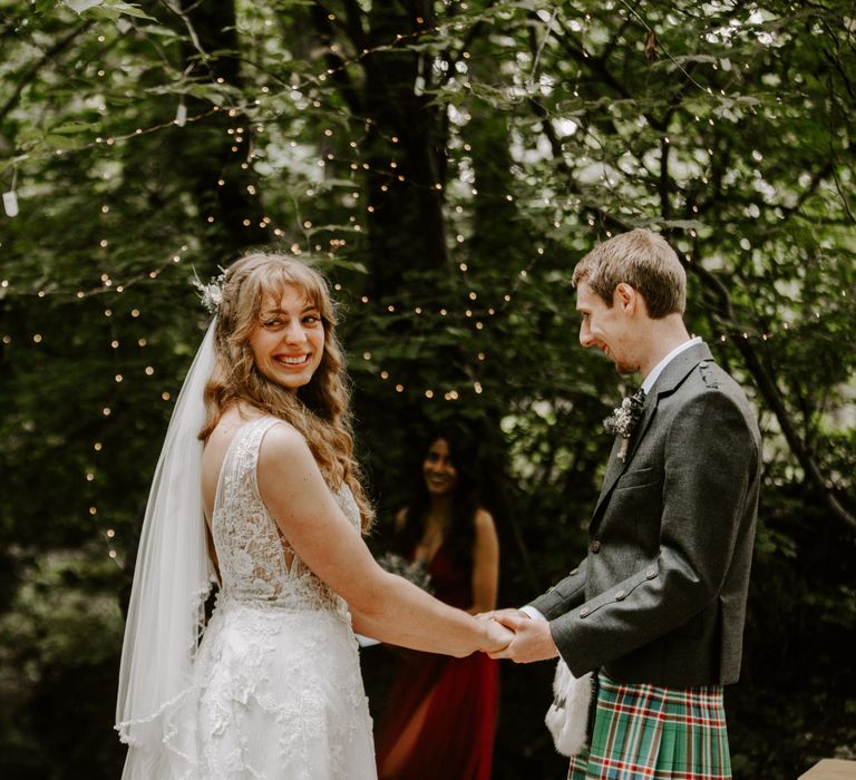 Bride in floral wedding gown and groom in traditional Scottish kilt marry in the Highlands amongst fairy lights