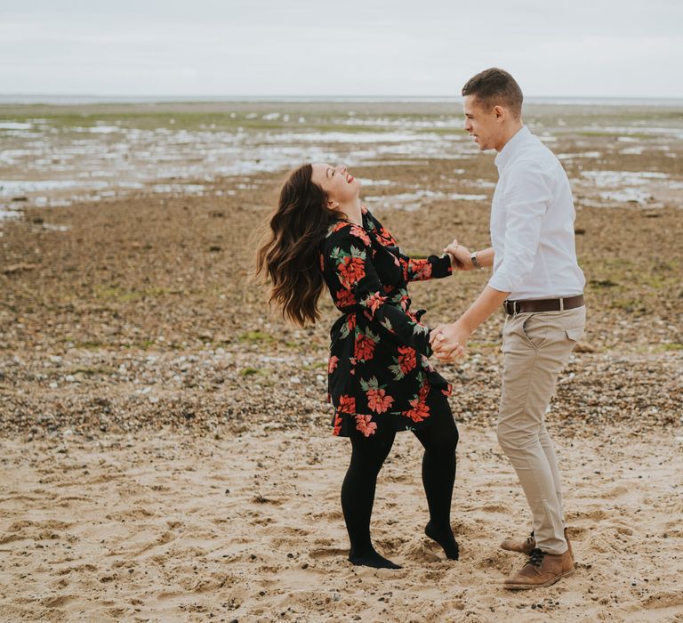 Bride-to-be with long brunette hair laughing on the beach in a black and red floral dress and tights with her groom-to-be in a white shirt and chino's 