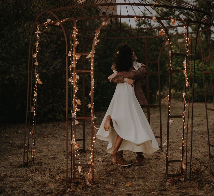 Groom picking up his bride in a wore framed pergola decorated with fairy lights 