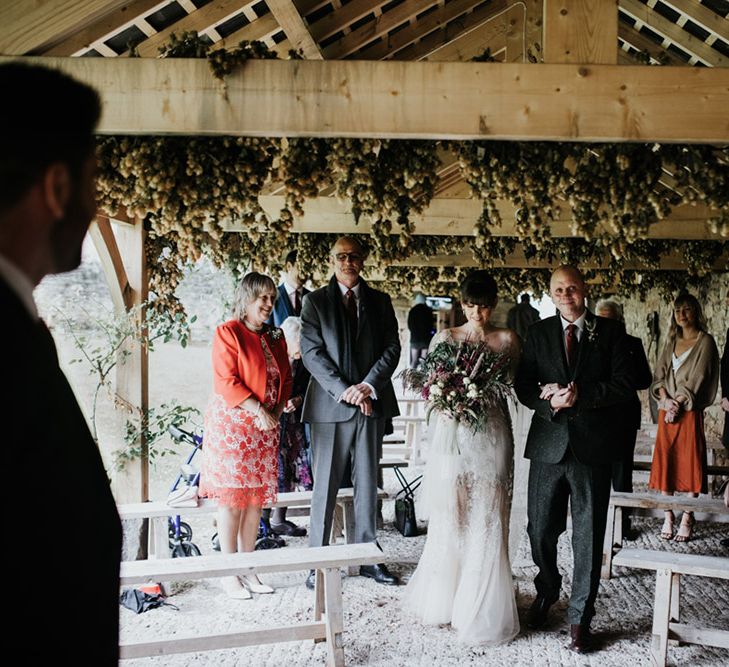 A bride is walked down the aisle by her dad. The venue is outdoor but under cover of beams and foliage. She wears a homemade wedding dress.