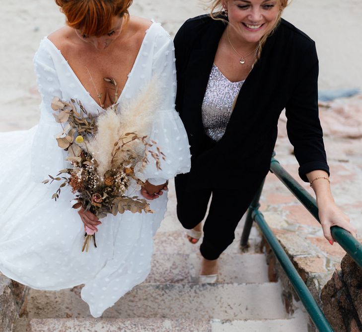 Two brides, one in dress holding bouquet and one in suit walking up beach steps