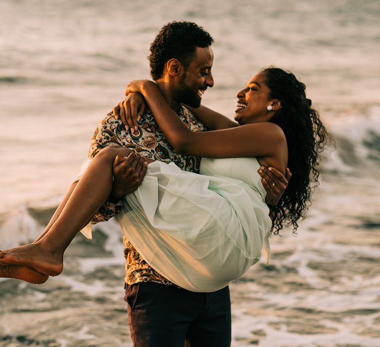 Groom carries bride along the beach as the ocean moves behind them