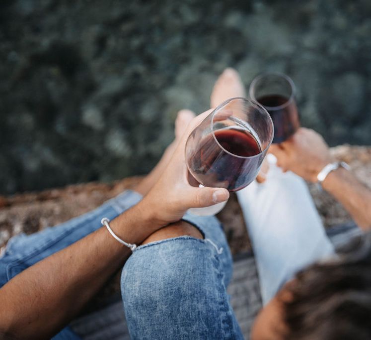 Grooms hold glasses of red wine and sit together on the beach