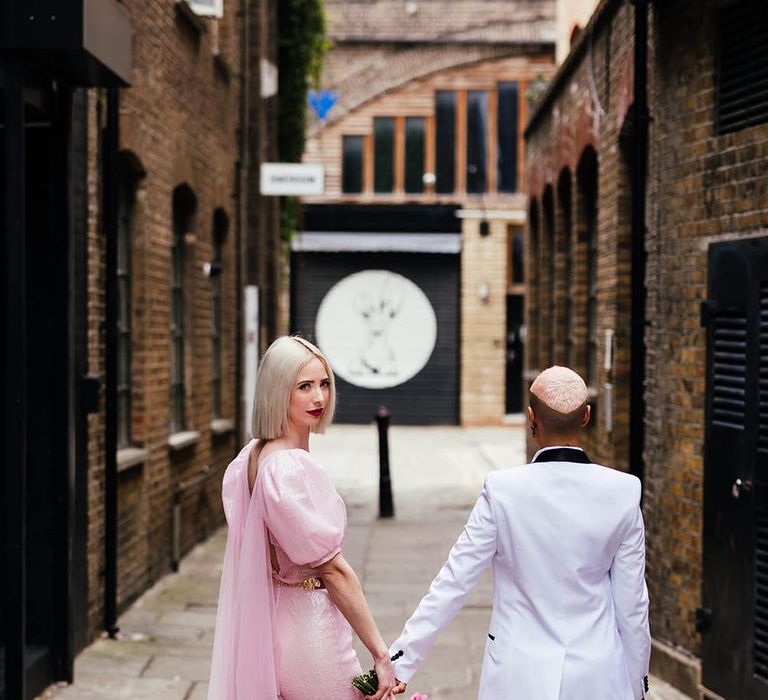 Bride in a fitted pink jumpsuit with a Watteau train and pink mules holding hands with her bride in a white tuxedo 