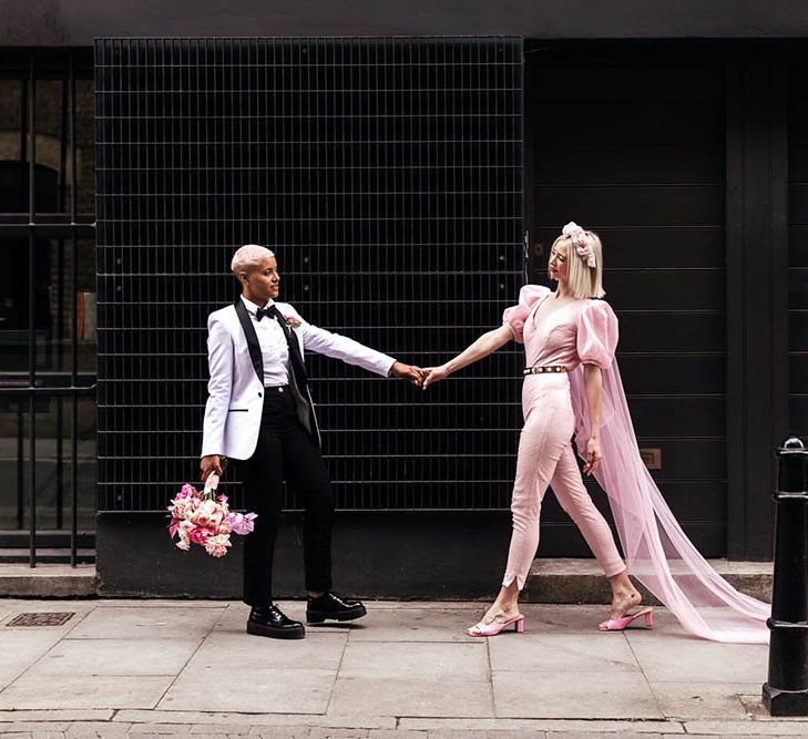 Bride in a white tuxedo jacket holding hands with her bride in a pink fitted jumpsuit with a Watteau train 