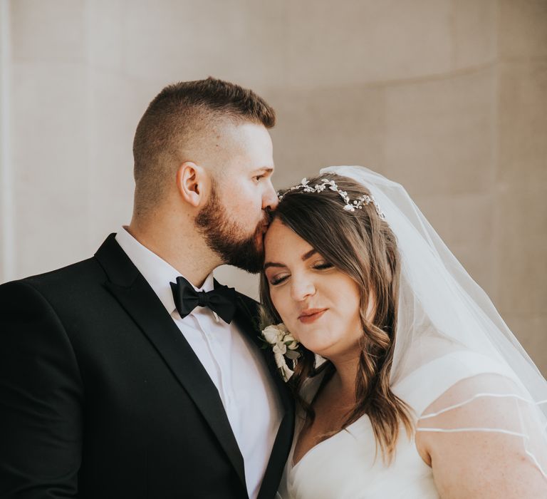 Groom kisses brides head on the morning of their wedding day as she holds classic white floral bouquet