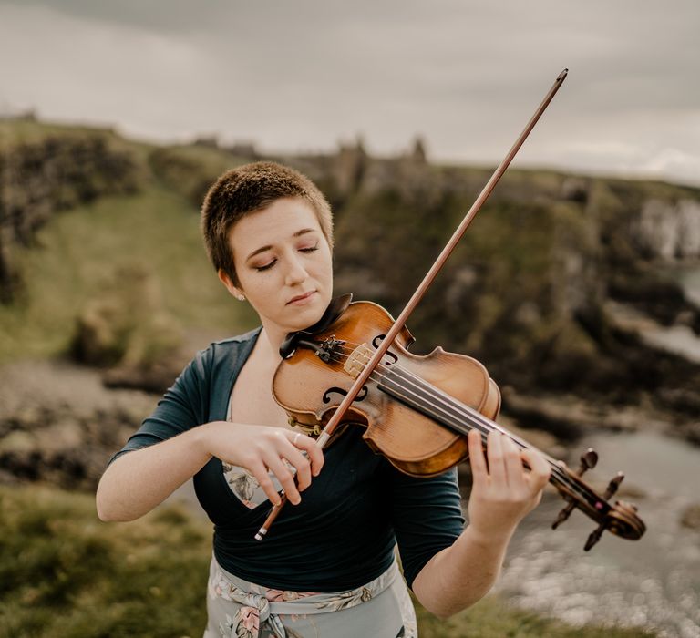 Violinist with short hair and blue wrap top plays with eyes closed at clifftop ceremony for Dunluce Castle wedding