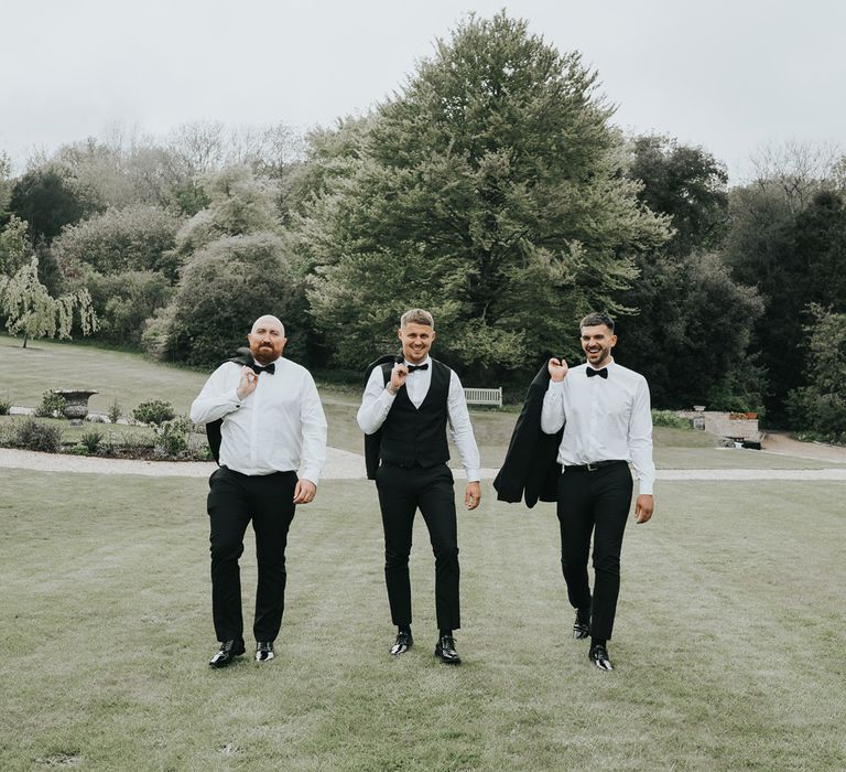 Groom and two groomsmen in black tuxedos walk across grounds at Came House Dorset with jackets slung over their shoulder and after wedding