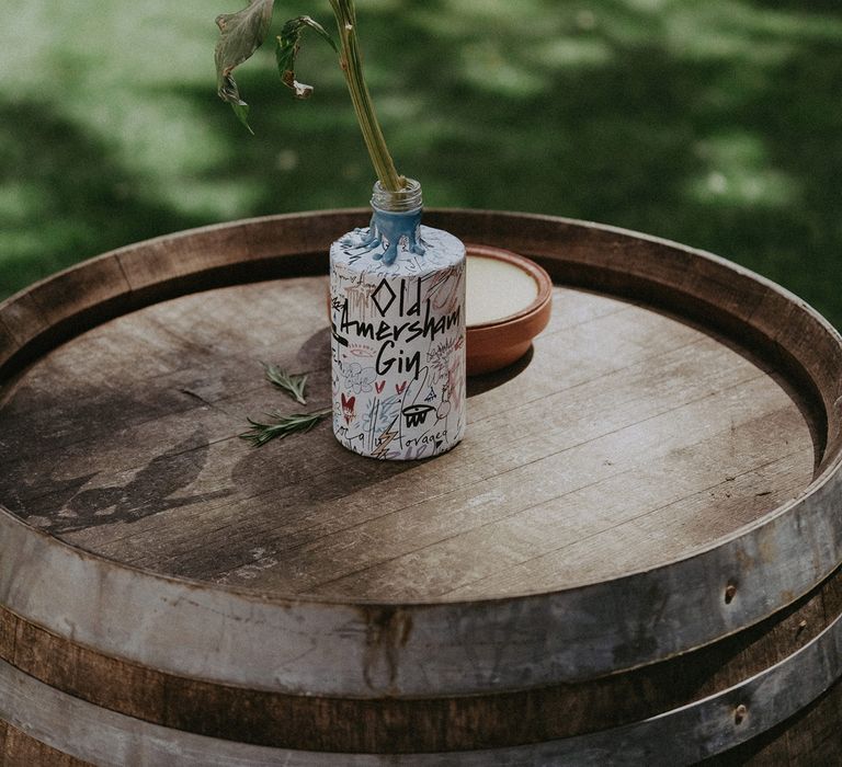 Multicoloured Old Amersham Gin bottle with pink flower on top of barrel for garden wedding reception 