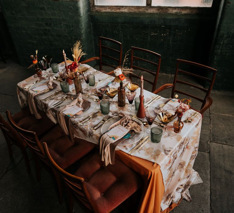 70s wedding tablescape with with patterned tablecloth, vases filled with flowers, coloured glassware and retro stationery 