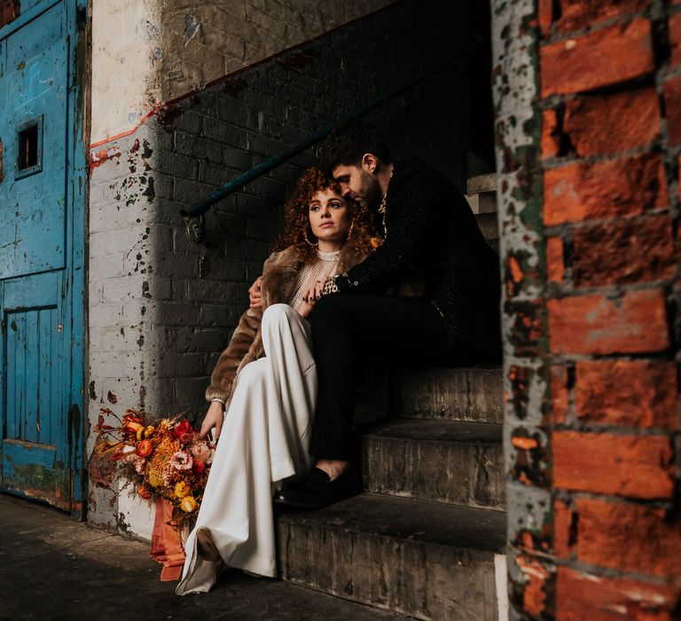 Bride and groom sitting on the steps in an industrial wedding venue in a jacquard suit jacket and faux fur coat 