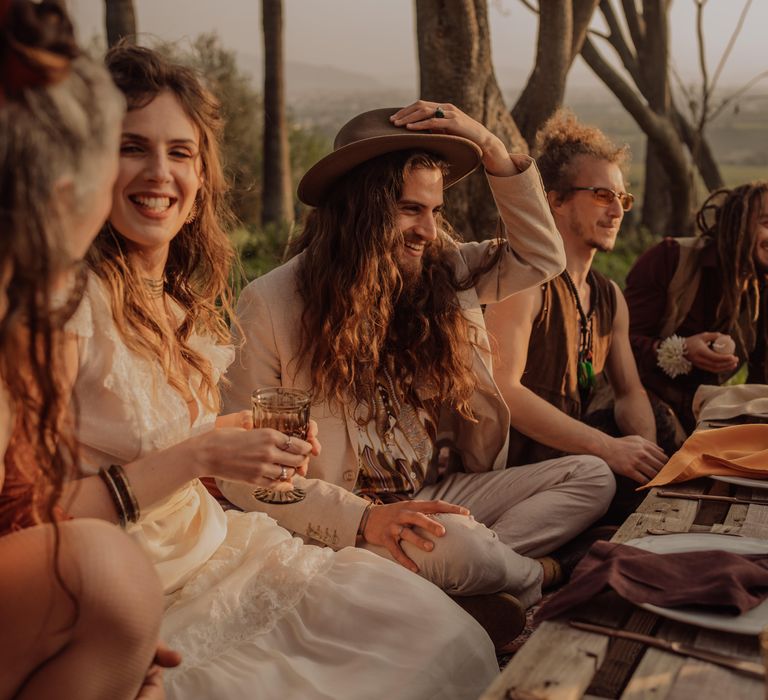 Wedding guests sit on the floor around rustic palette table setting for intimate reception in the outdoors of Sicily during sunset 