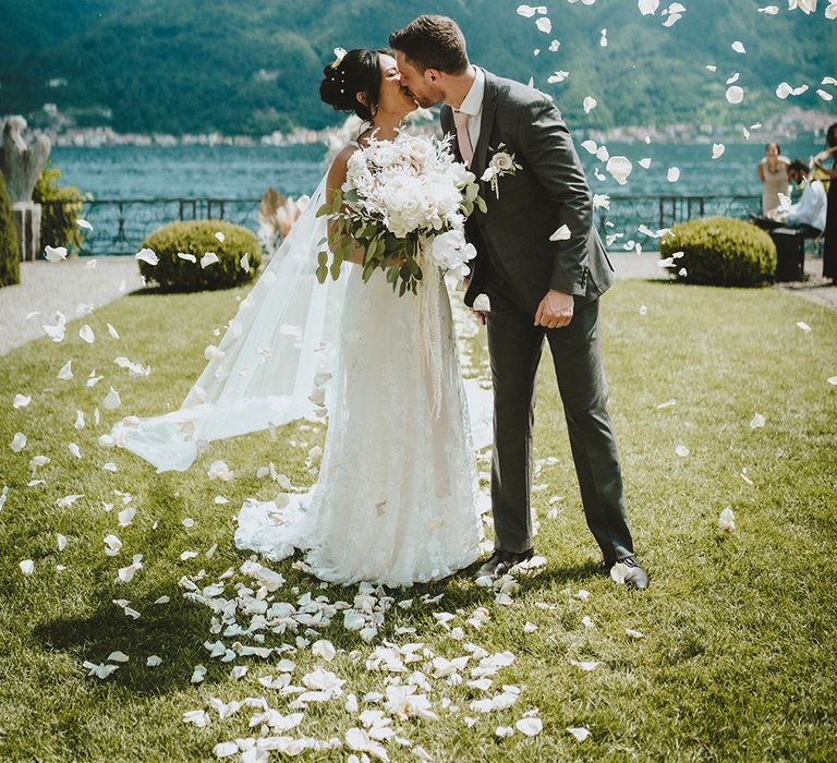 Bride & groom stand in front of Lake Como as white petals float beside them as they kiss one another after wedding ceremony