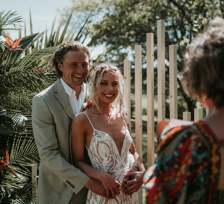 Groom in white shirt and linen suit has arms around front of bride in lace boho dress during outdoor wedding ceremony with tropical flowers for birds of paradise wedding inspiration