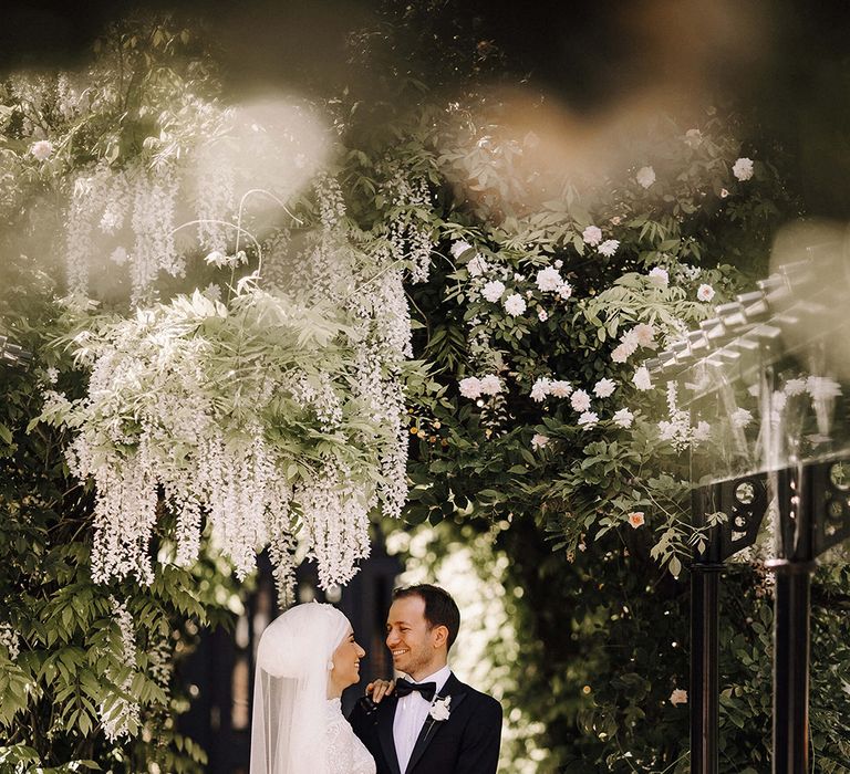 A Muslim bride stands with her new husband under an arch of white flowers. She wears all white and he wears black tie.