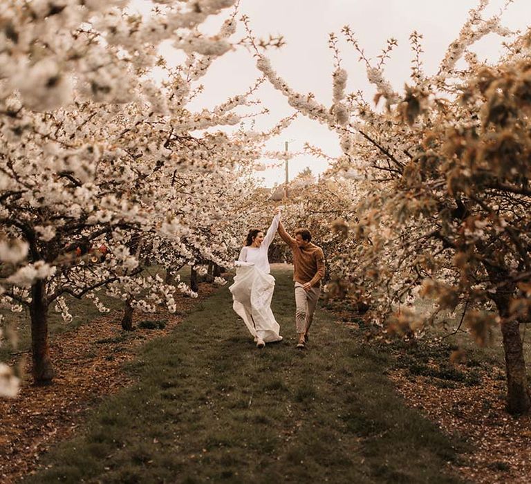 Bride and groom running through the trees in casual wedding attire during sunset 