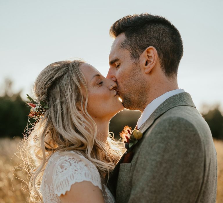 Bride in lace top open back wedding dress with blond curls and flowers in her hair hugs groom in grey herringbone suit in field at golden hour during late summer wedding at Wellington Wood Norfolk