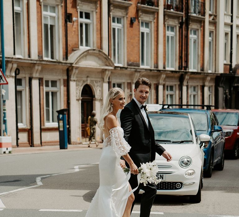 Stylish bride and groom in a satin and lace wedding dress with detachable sleeves and tuxedo crossing the road in Putney 